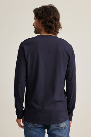 A man with shoulder-length hair faces away, wearing the ALVARO HENLEY by Velvet by Graham & Spencer over his jeans.