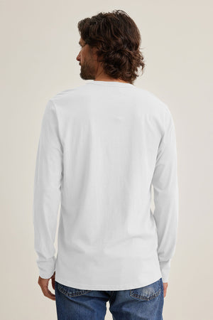 A man with dark hair, seen from the back, wears a white ALVARO HENLEY by Velvet by Graham & Spencer and blue jeans against a plain background, ideal for layering.