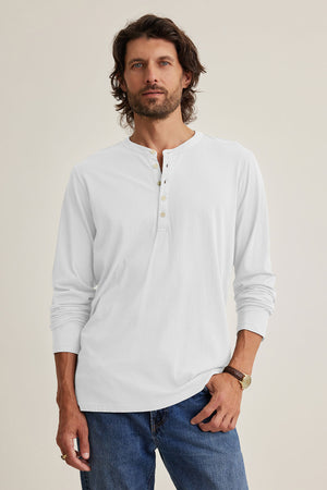 A man with shoulder-length hair stands against a plain background wearing Velvet by Graham & Spencer's ALVARO HENLEY in soft Pima jersey, paired with blue jeans.