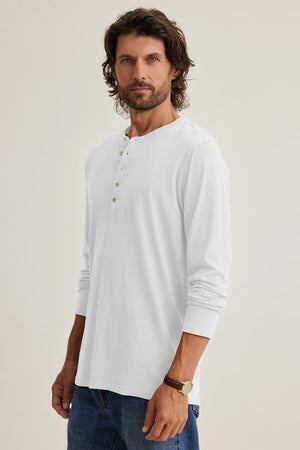 A bearded individual with long hair dons the ALVARO HENLEY by Velvet by Graham & Spencer, ideal for layering, combined with jeans and a watch, against a simple backdrop.