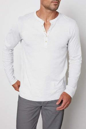 A man with a shaved head and short beard is wearing a white, long-sleeve ALVARO HENLEY shirt by Velvet by Graham & Spencer made from whisper knit fabric and gray pants. He stands with one hand on his hip, looking slightly to the side against a plain background.