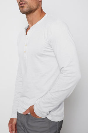 A man with a shaved head and a short beard wears a white long-sleeve ALVARO HENLEY by Velvet by Graham & Spencer and gray pants, standing against a plain white background.