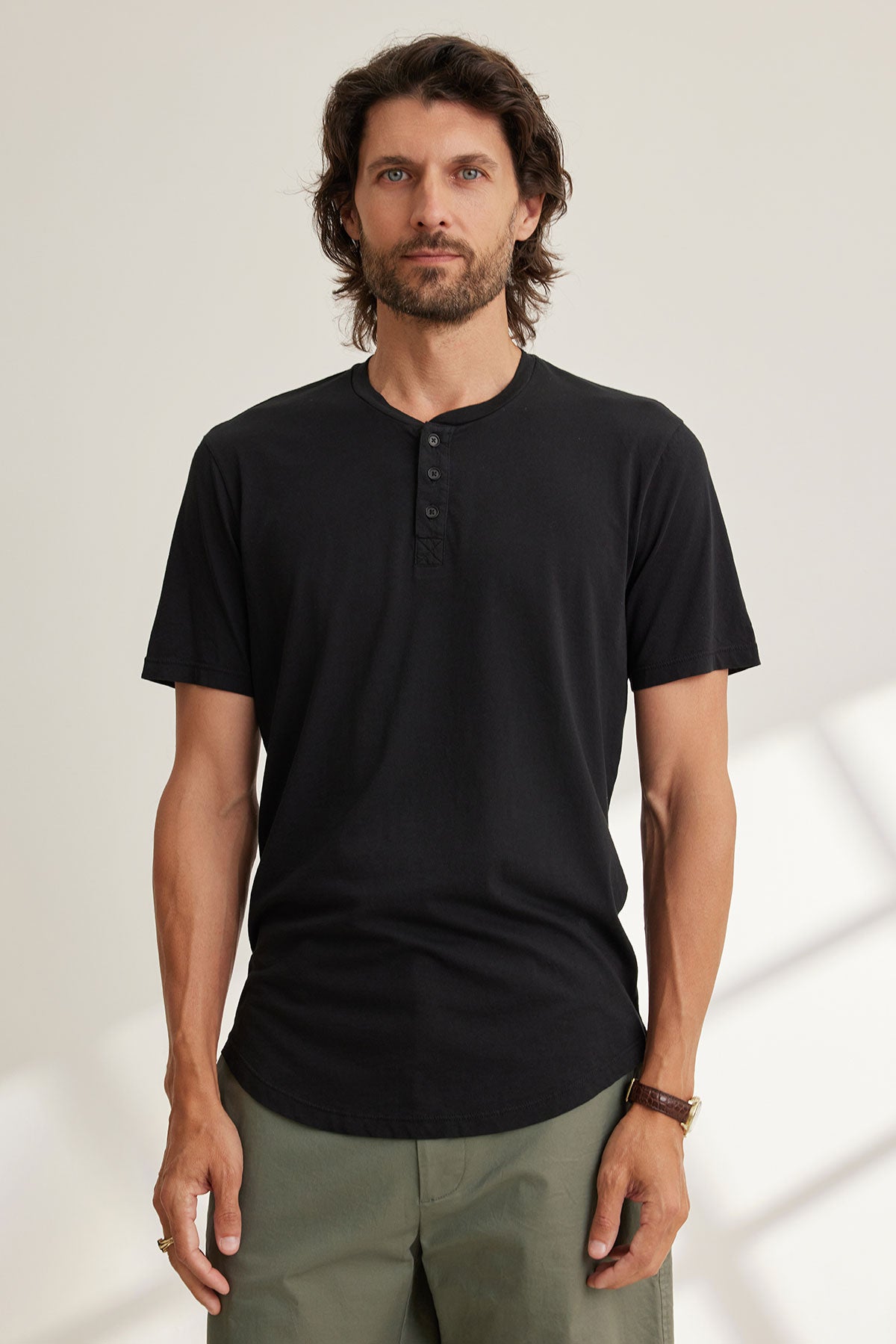 A man with long hair and a beard models Velvet by Graham & Spencer's FULTON HENLEY, a black 3-button shirt featuring a curved hemline, paired with green pants, against a light background.