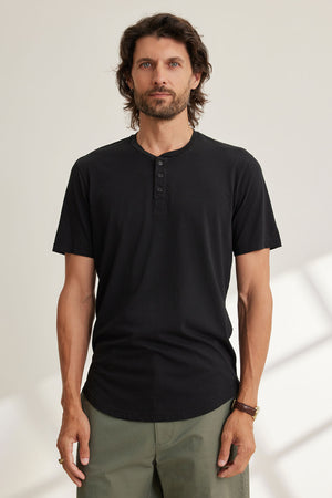 A man with long hair and a beard models Velvet by Graham & Spencer's FULTON HENLEY, a black 3-button shirt featuring a curved hemline, paired with green pants, against a light background.