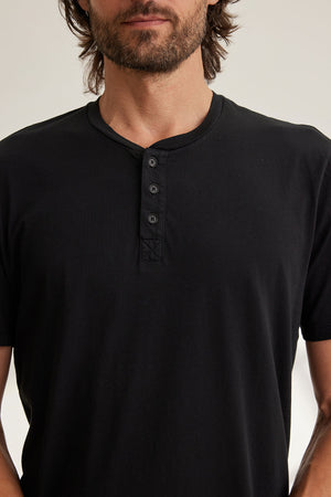 The image displays a person wearing the FULTON HENLEY by Velvet by Graham & Spencer, a black short-sleeve henley shirt with a curved hemline and four buttons, from shoulders to waist against a neutral background.