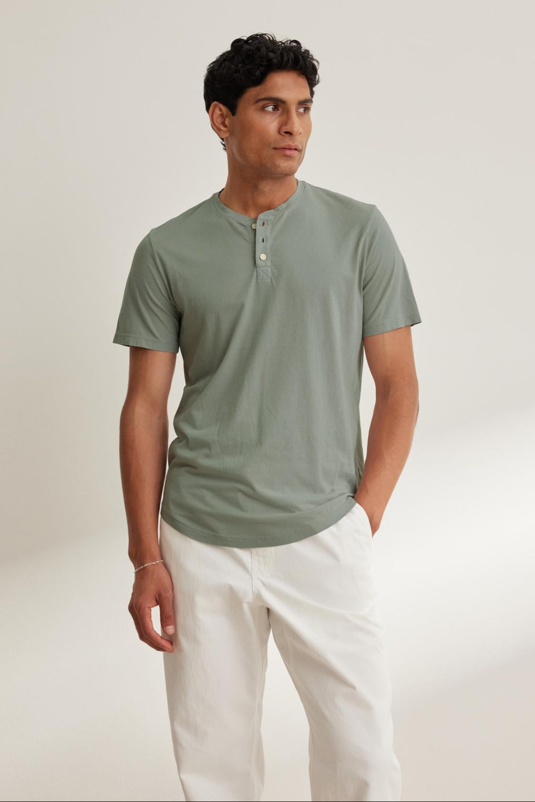 A person in a green Velvet by Graham & Spencer FULTON HENLEY short-sleeve tee made from lightweight Pima jersey and white pants stands against a plain light background.-38658659483841