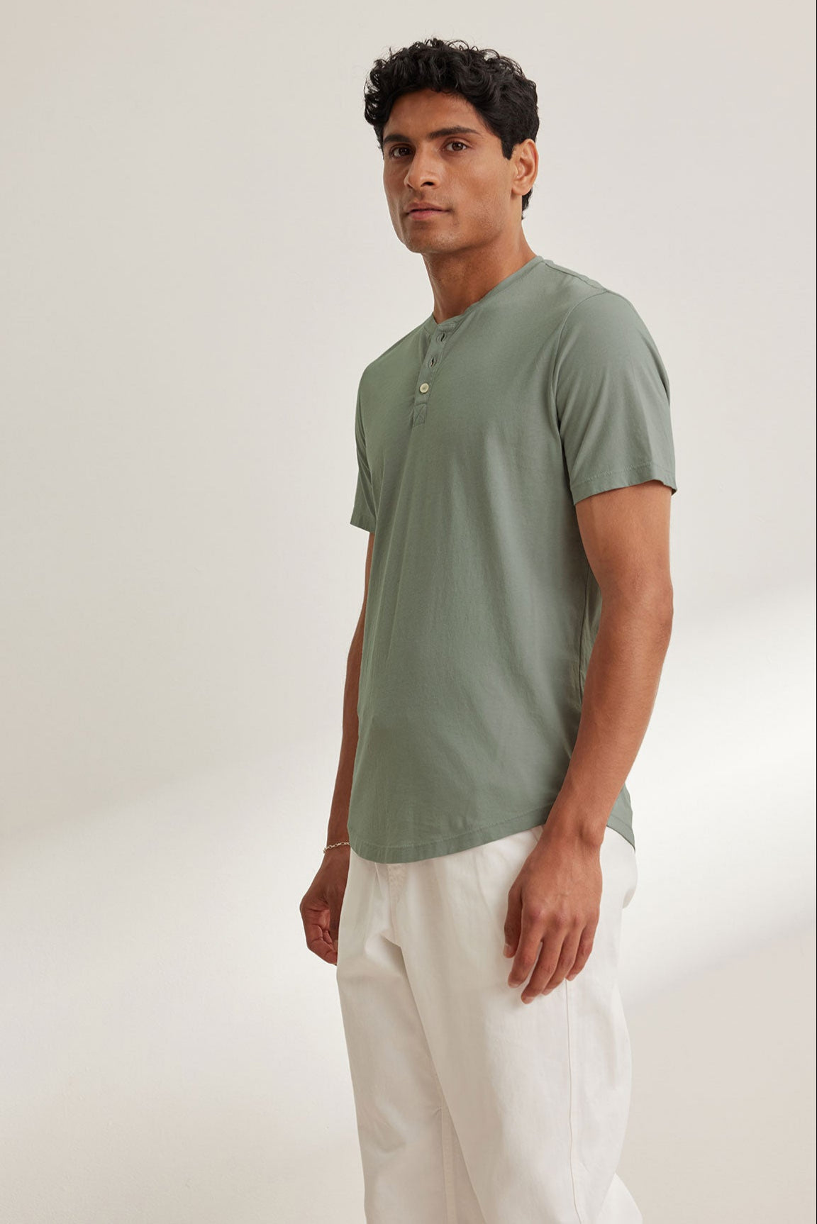 A person in a light green FULTON HENLEY by Velvet by Graham & Spencer with a curved hemline and white pants stands against a plain background.