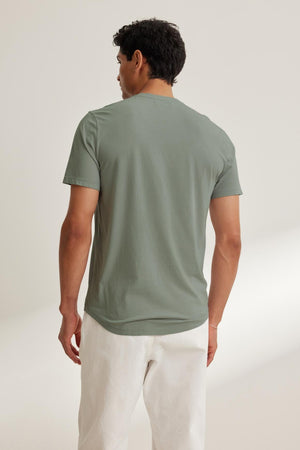 A man stands against a neutral background, wearing Velvet by Graham & Spencer's FULTON HENLEY in green, crafted from lightweight Pima jersey, paired with white pants.