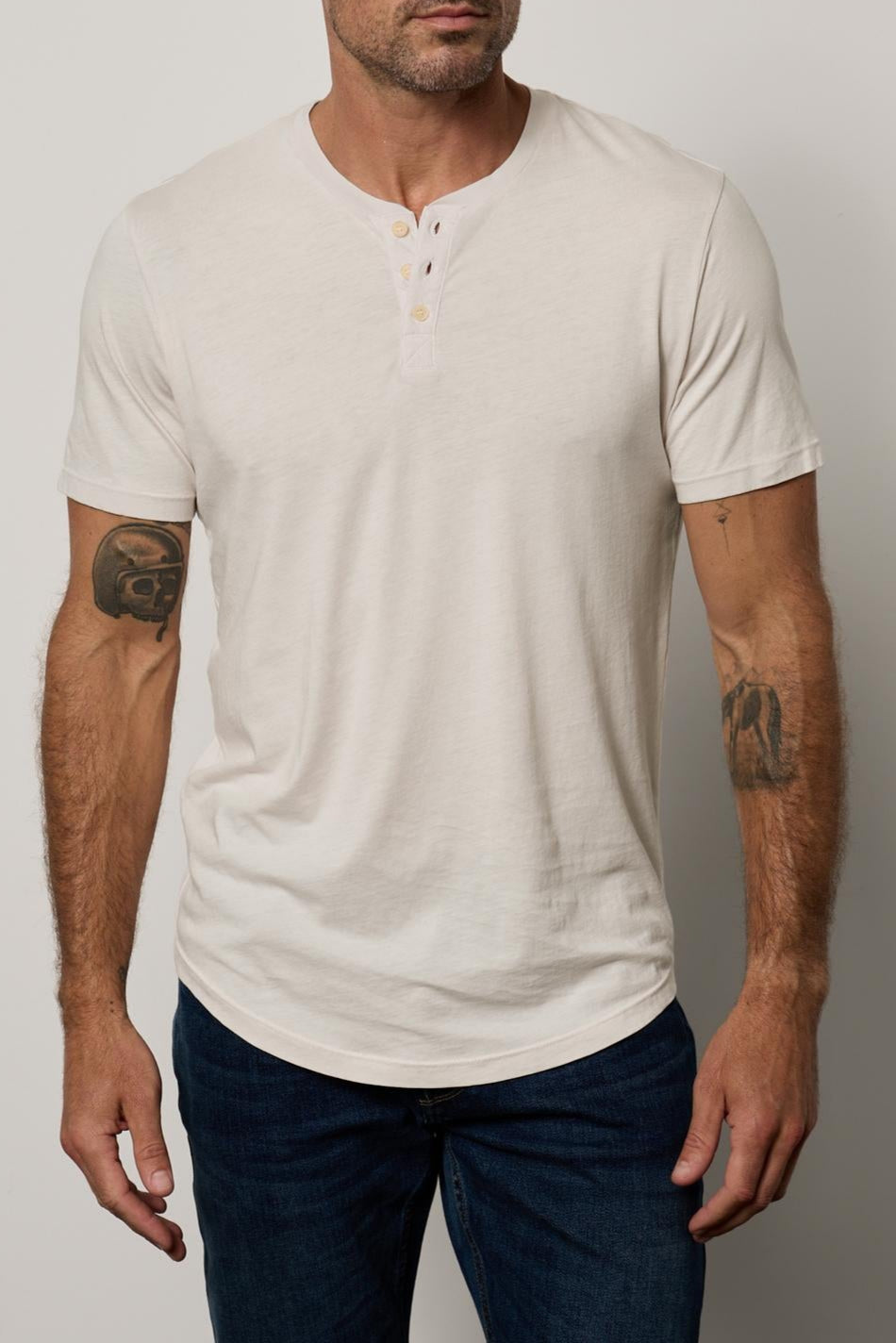 A bald man with tattoos on both arms, wearing a white Velvet by Graham & Spencer Fulton Henley with a curved hemline, standing against a neutral background.-36890721943745