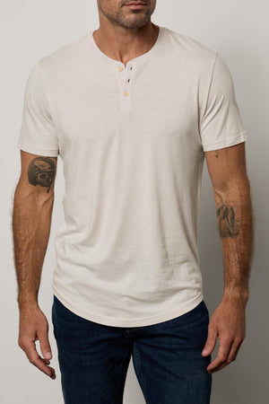 A bald man with tattoos on both arms, wearing a white Velvet by Graham & Spencer Fulton Henley with a curved hemline, standing against a neutral background.