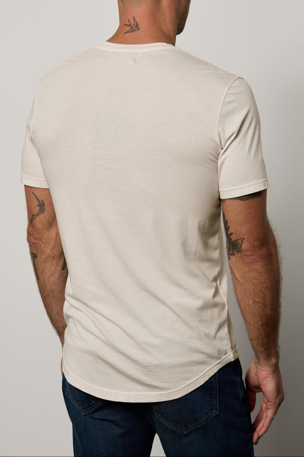 A man with a shaved head, wearing a beige, lightweight Velvet by Graham & Spencer FULTON HENLEY with a curved hemline and blue jeans, is shown from the back. He has visible tattoos on his arms and neck.-36890722009281