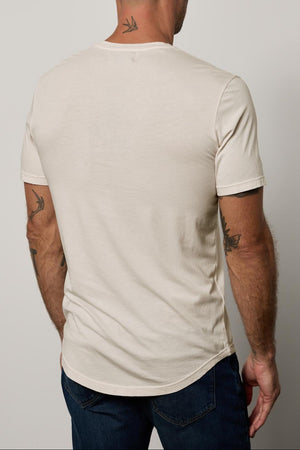 A man with a shaved head, wearing a beige, lightweight Velvet by Graham & Spencer FULTON HENLEY with a curved hemline and blue jeans, is shown from the back. He has visible tattoos on his arms and neck.