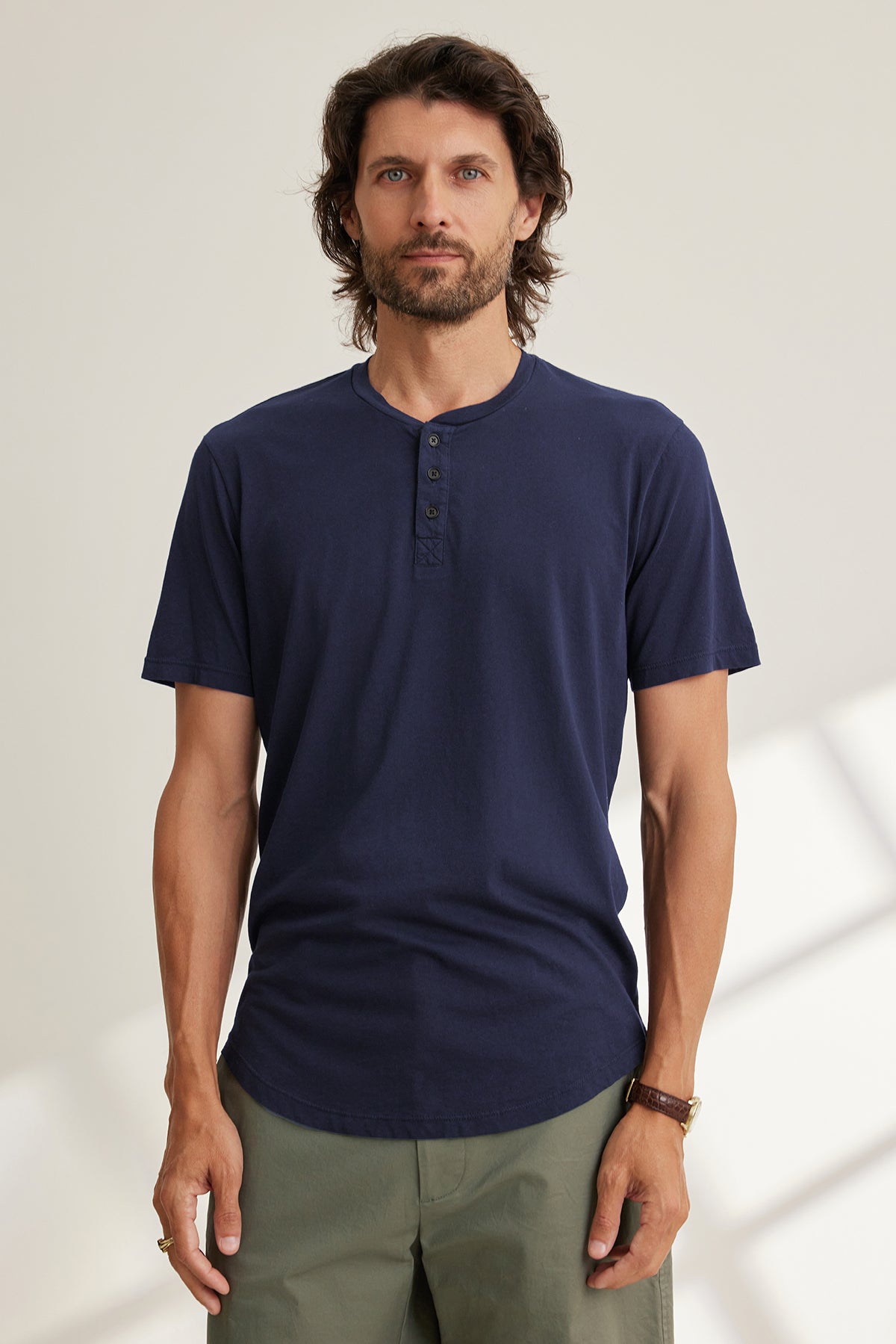 A man with long hair and a beard wears a navy FULTON HENLEY by Velvet by Graham & Spencer and green pants, standing against a light-colored background.