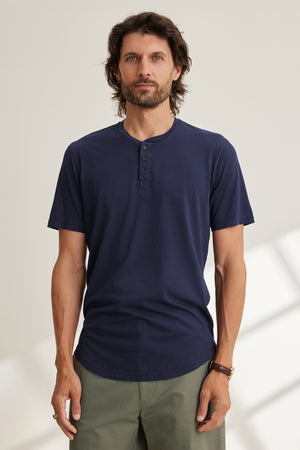 A man with long hair and a beard wears a navy FULTON HENLEY by Velvet by Graham & Spencer and green pants, standing against a light-colored background.