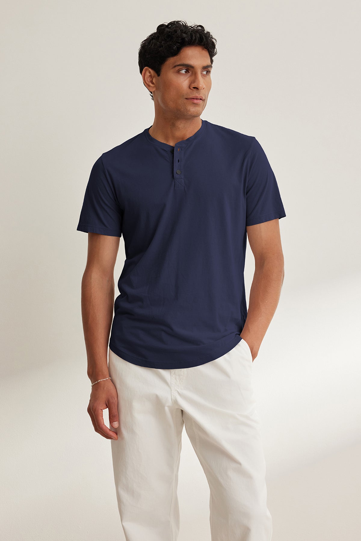 Against a plain backdrop, a person wears Velvet by Graham & Spencer's navy blue FULTON HENLEY short-sleeve Pima jersey tee with white pants, glancing to the side with one hand in their pocket.