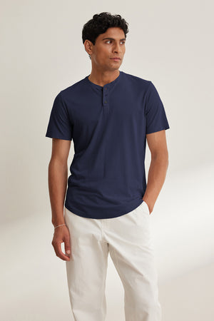 Against a plain backdrop, a person wears Velvet by Graham & Spencer's navy blue FULTON HENLEY short-sleeve Pima jersey tee with white pants, glancing to the side with one hand in their pocket.