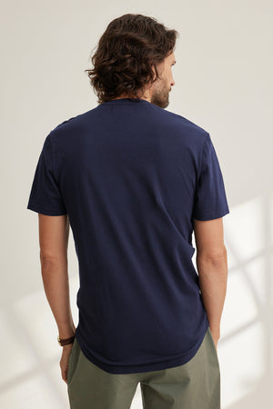 A man with curly hair stands with his back to the camera, wearing Velvet by Graham & Spencer's FULTON HENLEY lightweight navy T-shirt made from soft Pima jersey and olive green pants, set against a neutral background.