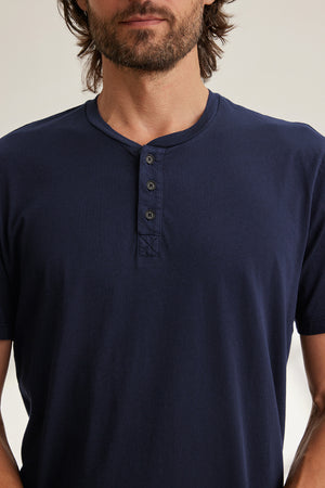A man stands against a neutral background wearing the Velvet by Graham & Spencer's navy blue FULTON HENLEY, a lightweight tee made from soft Pima jersey featuring three buttons.