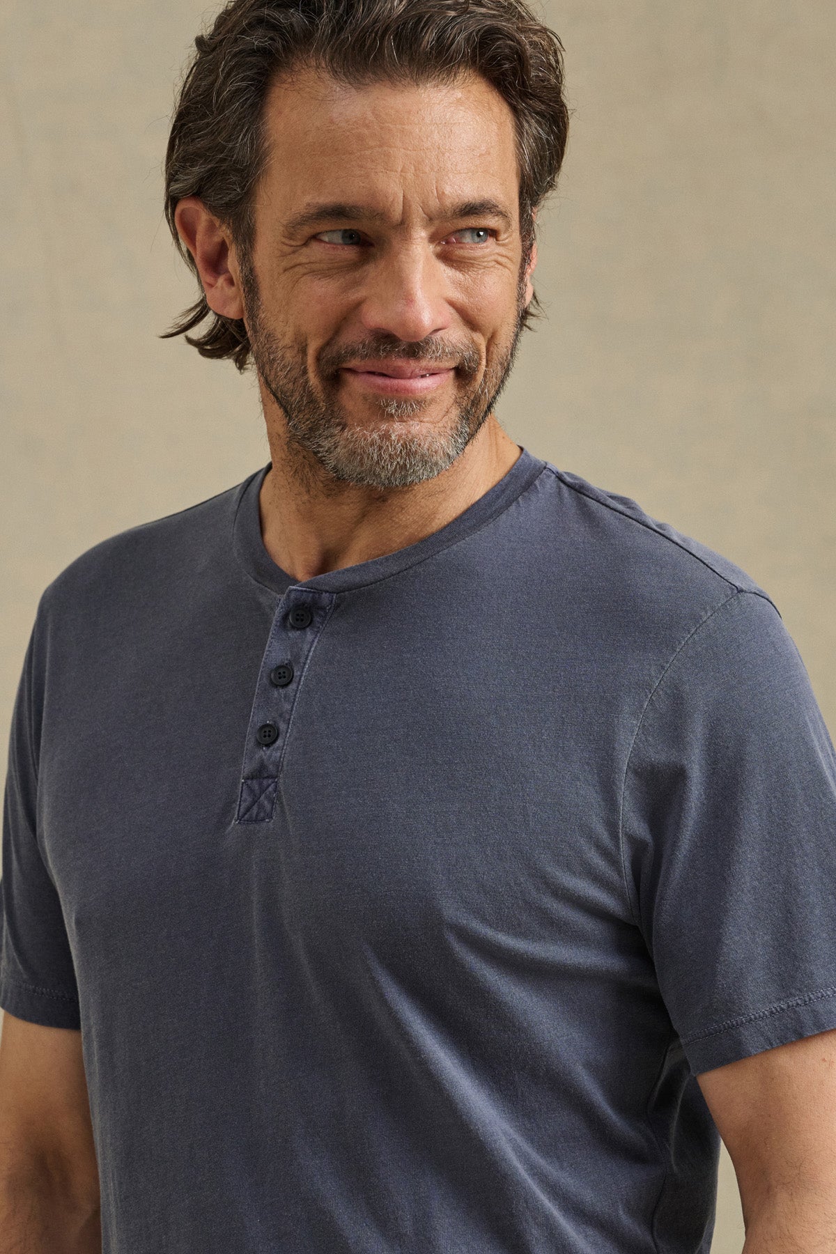 A middle-aged man with brown hair and a beard wears the FULTON HENLEY by Velvet by Graham & Spencer—a short-sleeve blue tee with a curved hemline—while looking to the side and smiling slightly.-41626555678913
