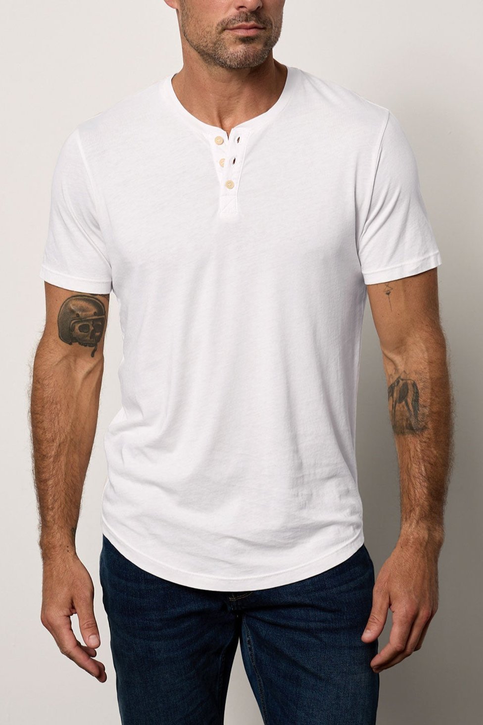 A man with a shaved head and tattoos on his arms, wearing a white Velvet by Graham & Spencer FULTON HENLEY tee, stands facing the camera with a neutral expression.-36640141115585