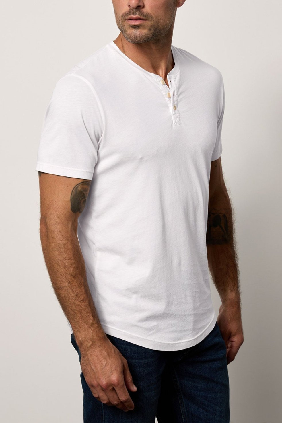 A man with a shaved head, wearing a white Velvet by Graham & Spencer Fulton Henley and dark jeans, stands against a plain background, looking to his left.-36640141148353