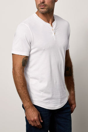 A man with a shaved head, wearing a white Velvet by Graham & Spencer Fulton Henley and dark jeans, stands against a plain background, looking to his left.