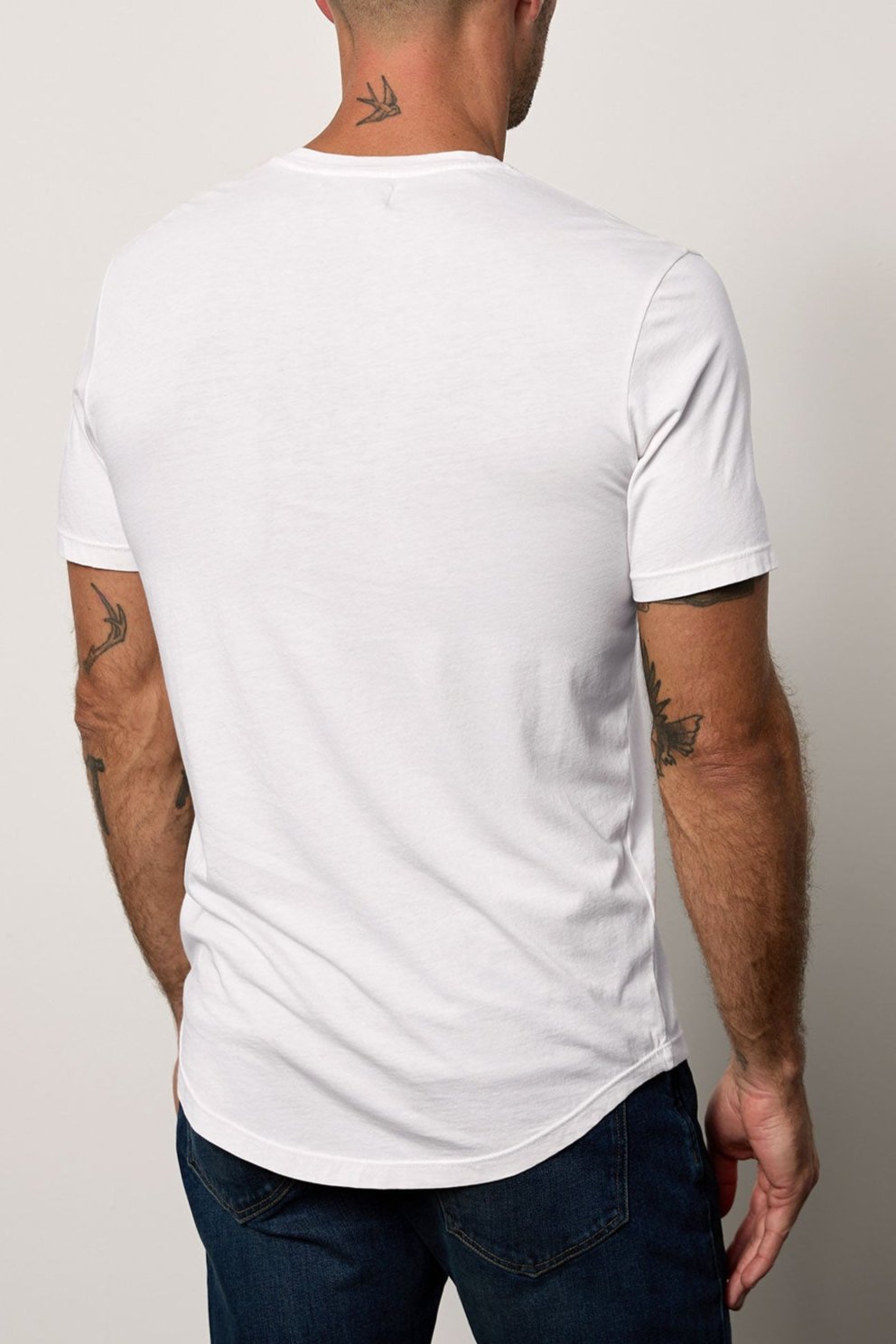 Man standing with his back to the camera, wearing a white Velvet by Graham & Spencer Fulton henley tee with a curved hemline, revealing tattoos on his arms and neck.-36640141181121