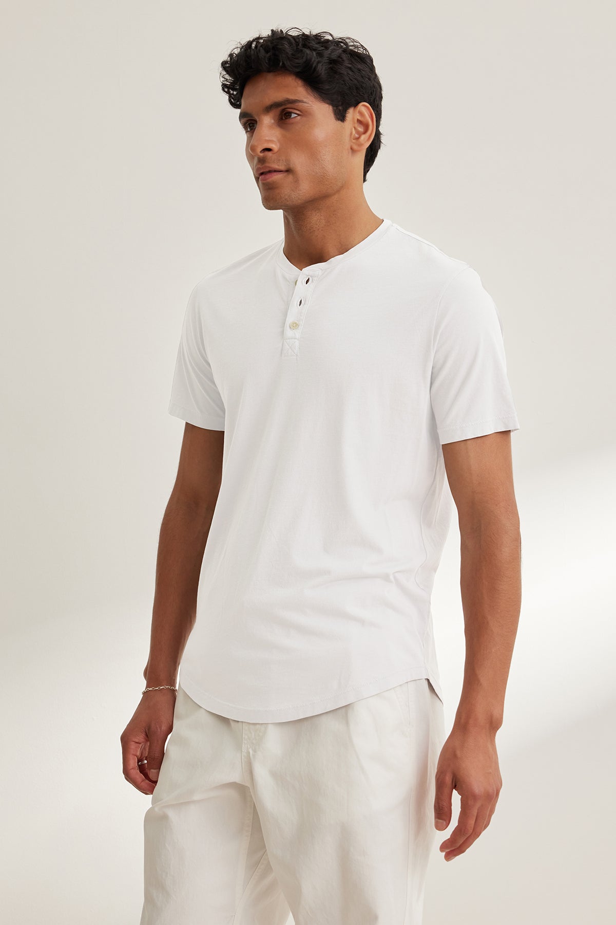 A person stands against a light background, wearing the FULTON HENLEY by Velvet by Graham & Spencer, a luxurious Pima jersey white short-sleeve tee, paired with matching white pants.-39029876162753