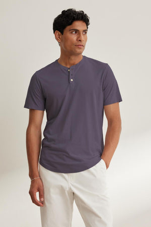 A man models the stylish curved hemline of the FULTON HENLEY by Velvet by Graham & Spencer, crafted from lightweight Pima jersey in dark gray, as he stands against a plain background with white pants.