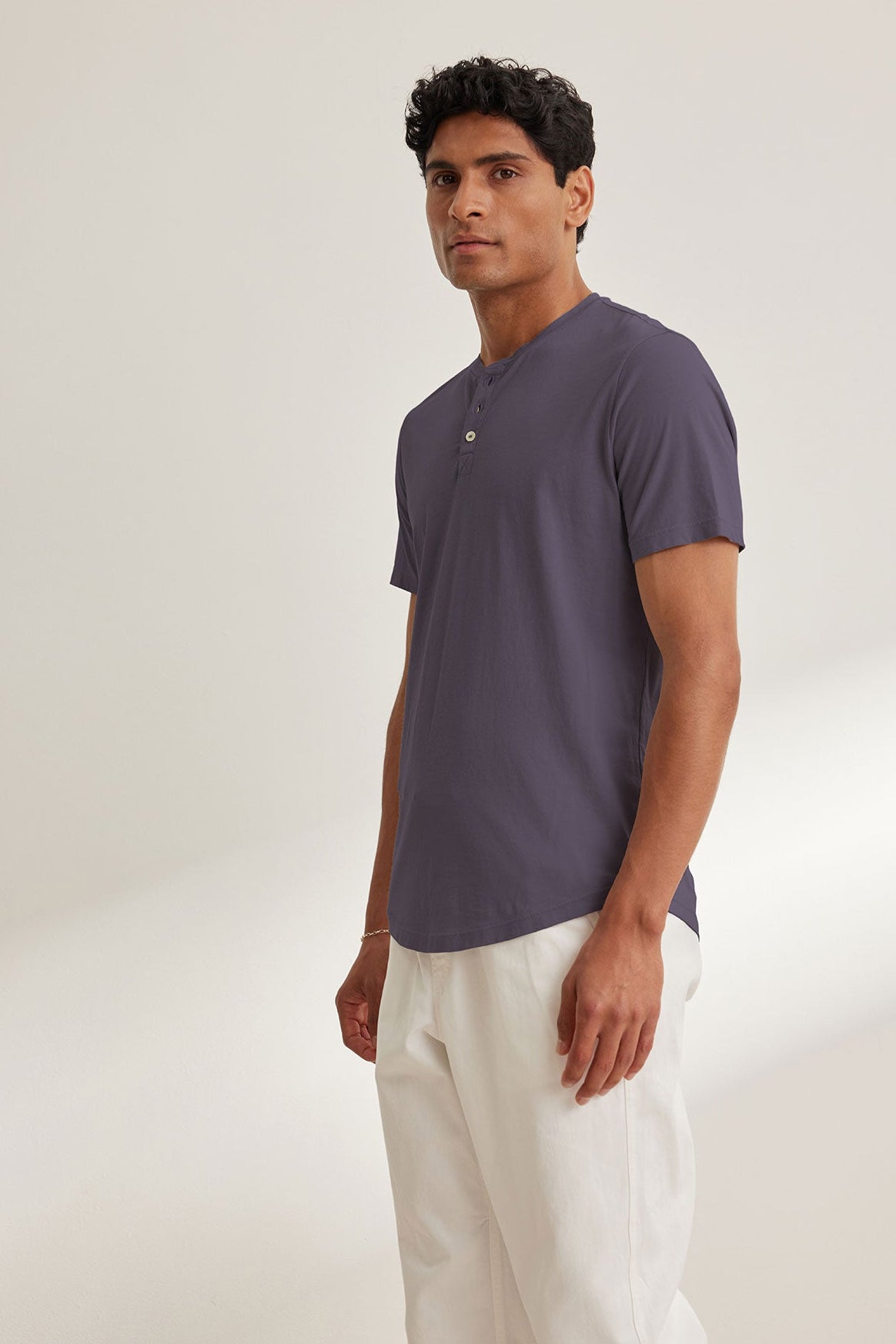 A person stands against a plain background wearing the Velvet by Graham & Spencer "FULTON HENLEY" in dark gray with a curved hemline and white pants.-38670204797121
