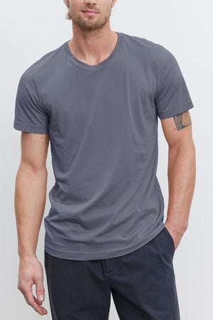 A person wearing a gray HOWARD TEE made of lightweight cotton knit from Velvet by Graham & Spencer and dark pants stands against a plain white background. One arm is resting by their side while the other is partially in their pocket, creating a relaxed yet perfect fit with a hint of vintage feel.