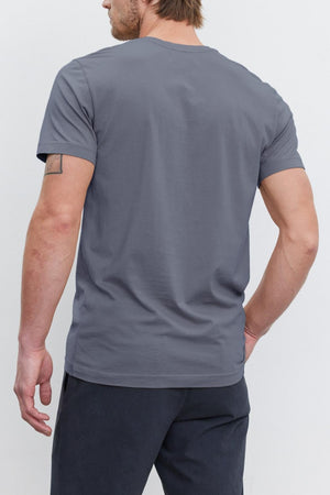 Person with brown hair and a beard, facing away from the camera, wearing a Velvet by Graham & Spencer HOWARD TEE with a vintage feel and dark pants.