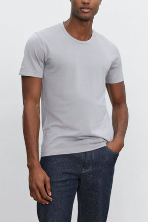 A person stands against a plain background, wearing a light gray HOWARD TEE from Velvet by Graham & Spencer made from lightweight cotton knit and dark blue jeans that boast the perfect fit.