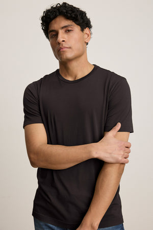 A person with short curly hair in a Velvet by Graham & Spencer HOWARD TEE, crafted from lightweight Pima jersey, and blue jeans stands against a plain background, facing forward with one arm crossed.