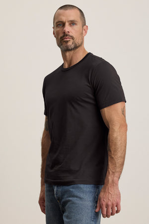 A bearded man wearing a perfectly fitting black HOWARD TEE by Velvet by Graham & Spencer and blue jeans stands against a plain background, exuding a vintage feel.