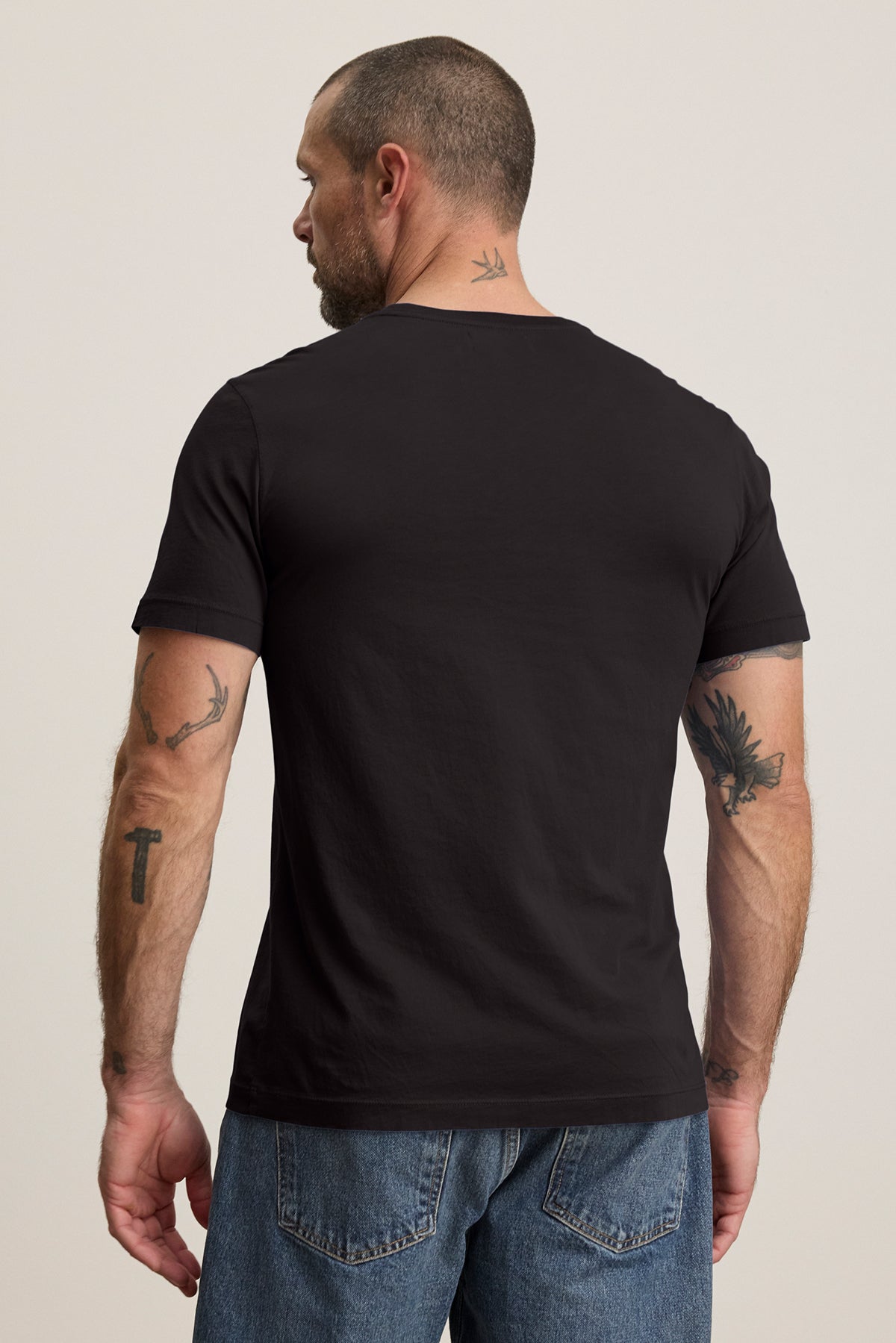 A tattooed individual wearing the HOWARD TEE, a black t-shirt by Velvet by Graham & Spencer made from lightweight Pima jersey, along with blue jeans is seen facing away from the camera.-39029790245057