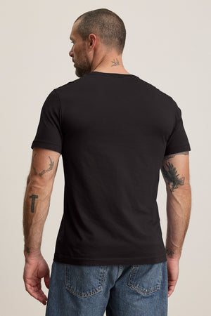 A tattooed individual wearing the HOWARD TEE, a black t-shirt by Velvet by Graham & Spencer made from lightweight Pima jersey, along with blue jeans is seen facing away from the camera.