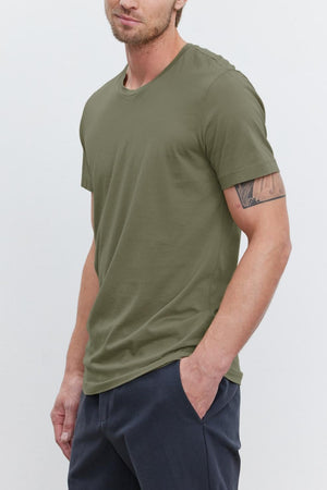 A young man with light brown hair wearing an olive green HOWARD TEE from Velvet by Graham & Spencer made from lightweight cotton knit and dark pants stands with his hands in his pockets against a plain white background, giving off a perfect fit.