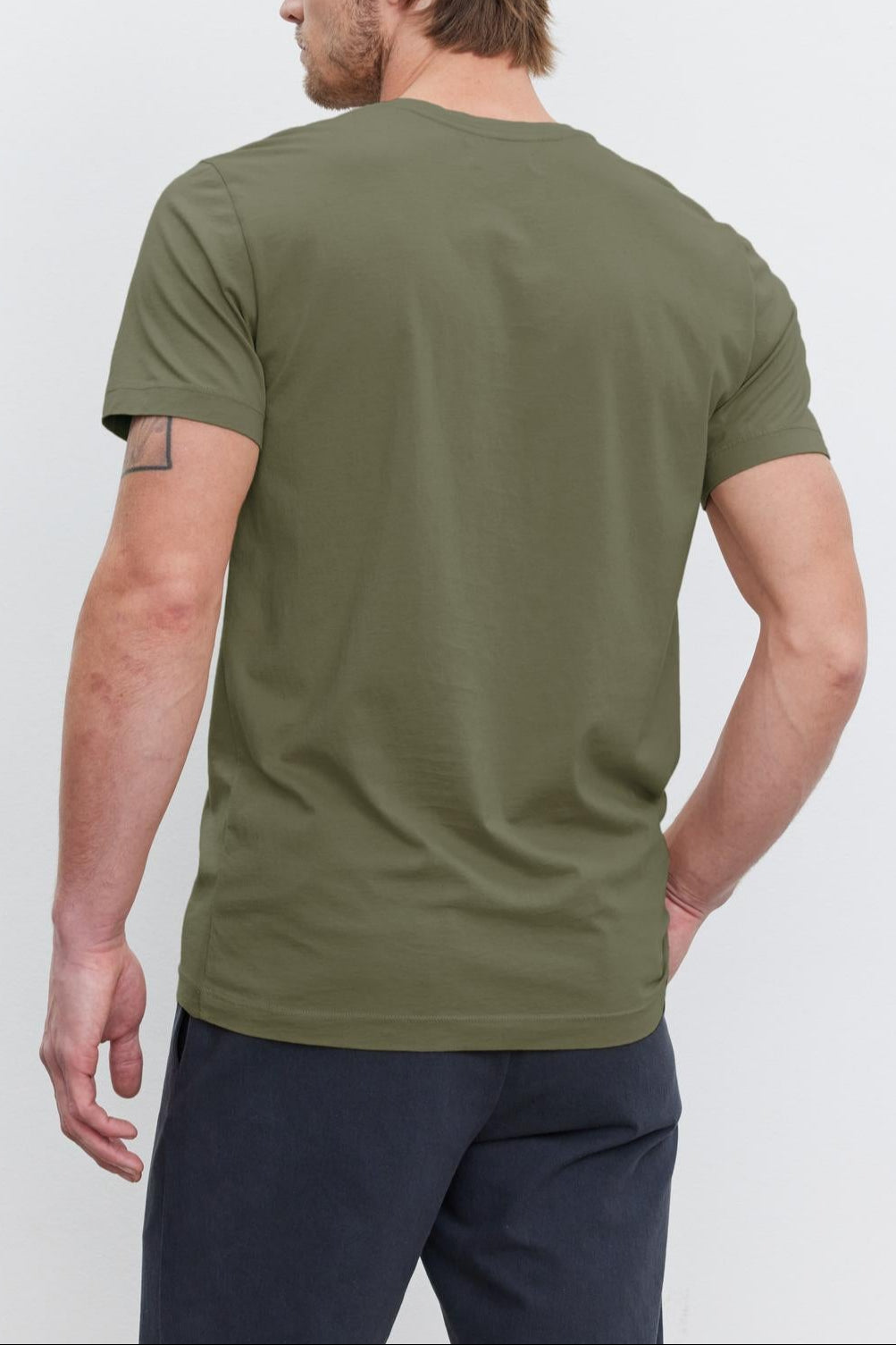 A person with brown hair and a beard, wearing a Velvet by Graham & Spencer HOWARD TEE with a vintage feel and dark pants, stands facing away from the camera, their right hand resting on their back.-37386103488705