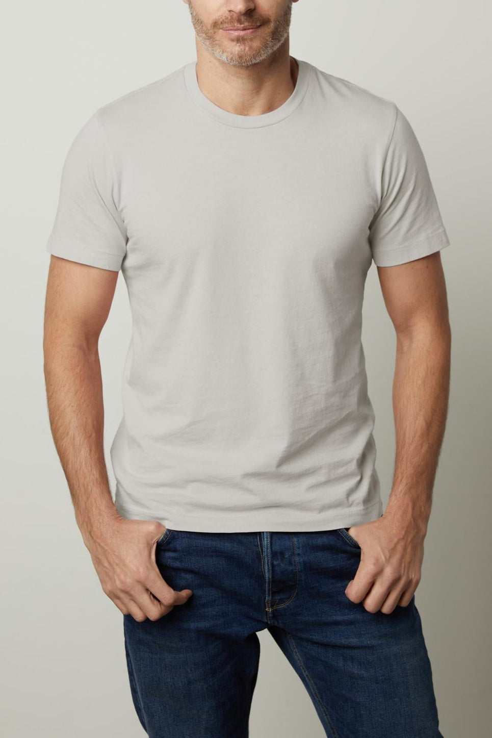 A man with short, dark hair and a beard wearing the light gray Velvet by Graham & Spencer HOWARD TEE and blue jeans stands with his hands in his pockets, exuding a vintage feel against a plain background.-37671772324033
