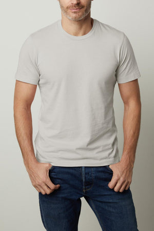 A man with short, dark hair and a beard wearing the light gray Velvet by Graham & Spencer HOWARD TEE and blue jeans stands with his hands in his pockets, exuding a vintage feel against a plain background.
