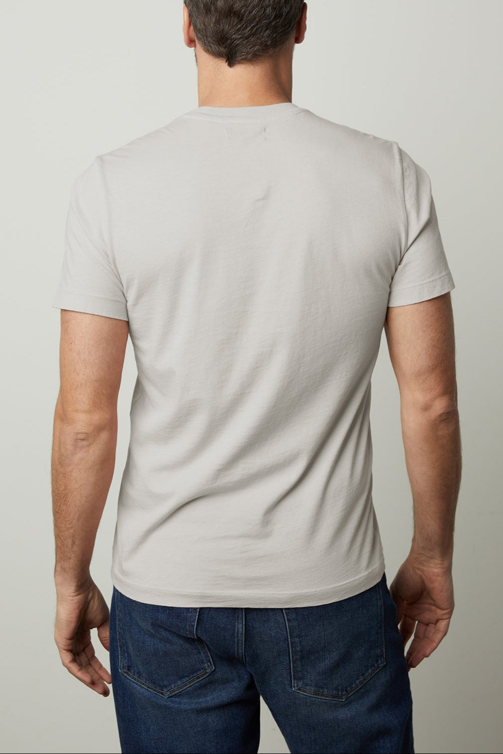 A man with short, dark hair stands with his back to the camera, wearing a light gray Velvet by Graham & Spencer HOWARD TEE made from lightweight cotton knit and dark blue jeans, offering a perfect fit.-37229772734657