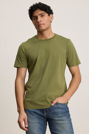 A man wears the HOWARD TEE by Velvet by Graham & Spencer, a plain olive green lightweight Pima jersey t-shirt, with blue jeans, standing against a neutral background with one hand in his pocket.