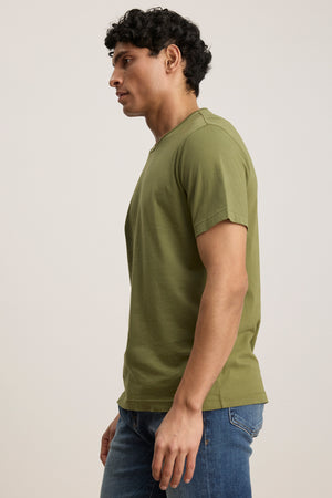 A person wears the HOWARD TEE by Velvet by Graham & Spencer, a short-sleeved olive green Pima jersey t-shirt, with blue jeans, standing left against a plain background.