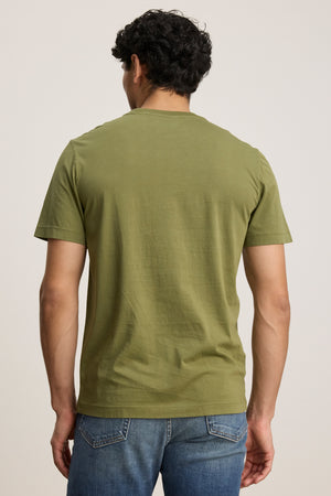 A man stands with his back to the camera, wearing the olive green HOWARD TEE by Velvet by Graham & Spencer, a lightweight Pima jersey short-sleeve shirt with a vintage feel, paired with blue jeans against a plain background.