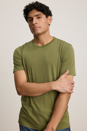 A man with short curly hair stands against a neutral background, wearing the HOWARD TEE by Velvet by Graham & Spencer—a plain olive green t-shirt made from lightweight Pima jersey—while crossing one arm over his body.