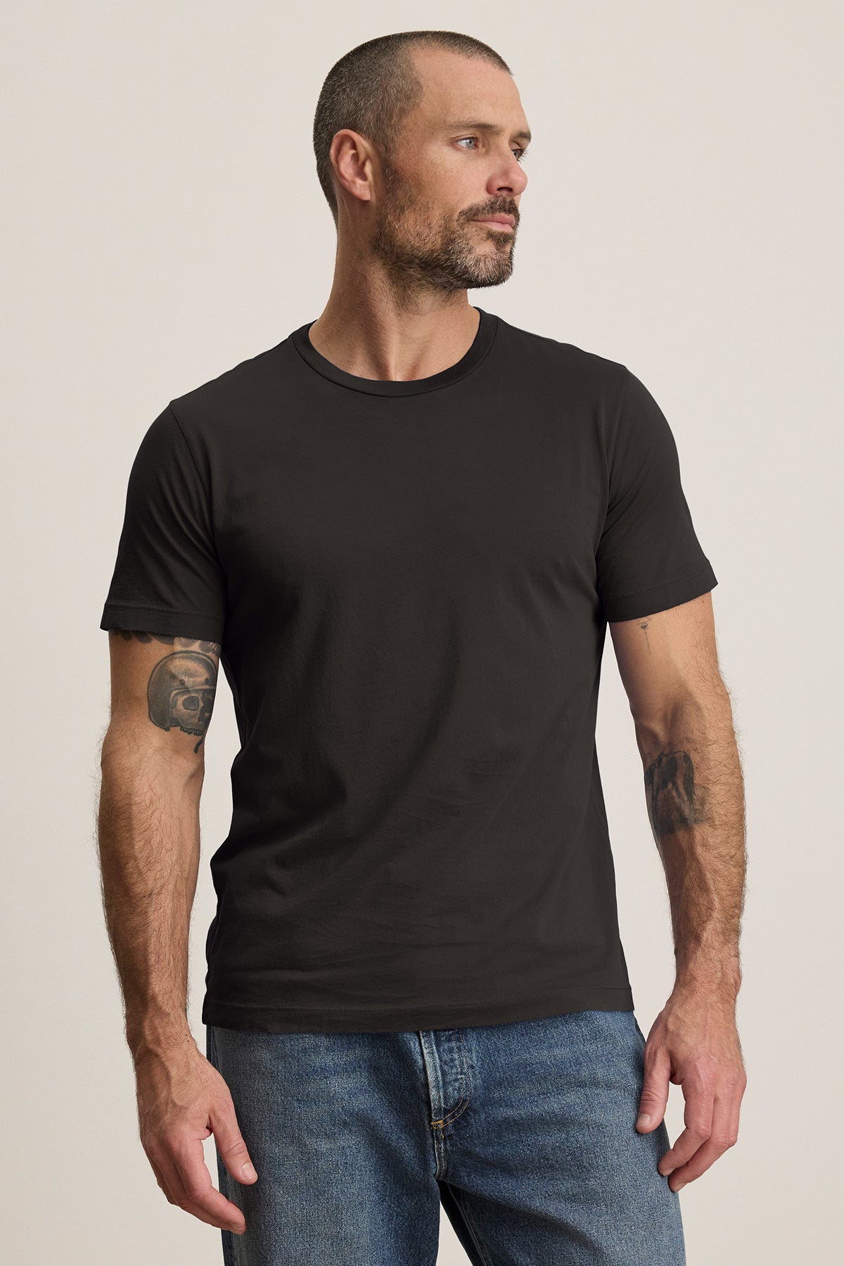 A bearded man effortlessly exudes style in jeans and a plain black HOWARD TEE from Velvet by Graham & Spencer. The lightweight Pima jersey provides a perfect fit as he stands confidently, embodying a vintage feel while gazing to the side.-39029851521217