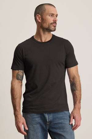 A bearded man effortlessly exudes style in jeans and a plain black HOWARD TEE from Velvet by Graham & Spencer. The lightweight Pima jersey provides a perfect fit as he stands confidently, embodying a vintage feel while gazing to the side.