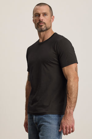 A bearded man in a black Velvet by Graham & Spencer HOWARD TEE, paired with blue jeans, faces the camera against a plain background. The tee is made from lightweight Pima jersey for comfort and an ideal fit.