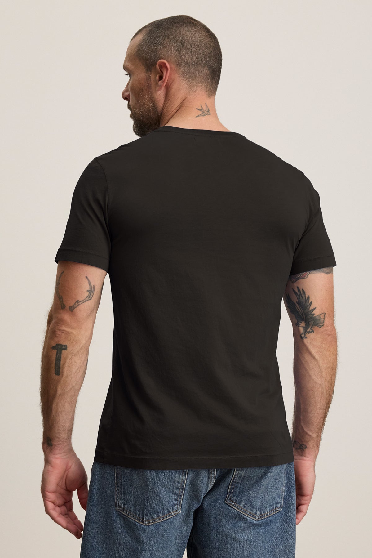 A tattooed man faces away, wearing the HOWARD TEE by Velvet by Graham & Spencer, a black t-shirt made from lightweight Pima jersey that offers a perfect fit, paired with blue jeans.